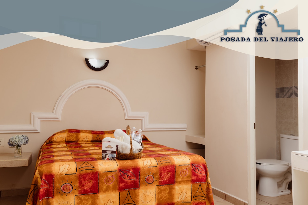 A comfortable room features a large bed dressed in a multicolored patterned blanket. A nightstand accompanies the bed, with toiletries arranged neatly nearby. The space is softly lit, highlighting the warm tones of the walls and the simple decor. A private bathroom is visible in the background.