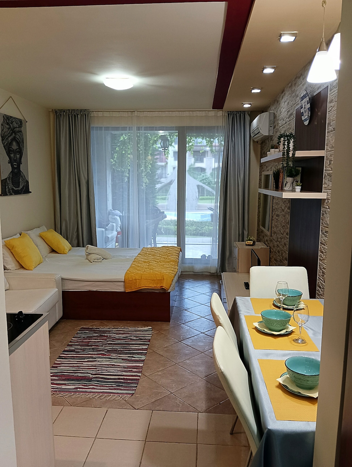 A bright studio apartment features a spacious sleeping area with a light-colored bedspread paired with vibrant yellow pillows. A dining table is set for four, accompanied by turquoise dishware. Large glass doors provide access to a patio area with greenery, enhancing the indoor ambiance.