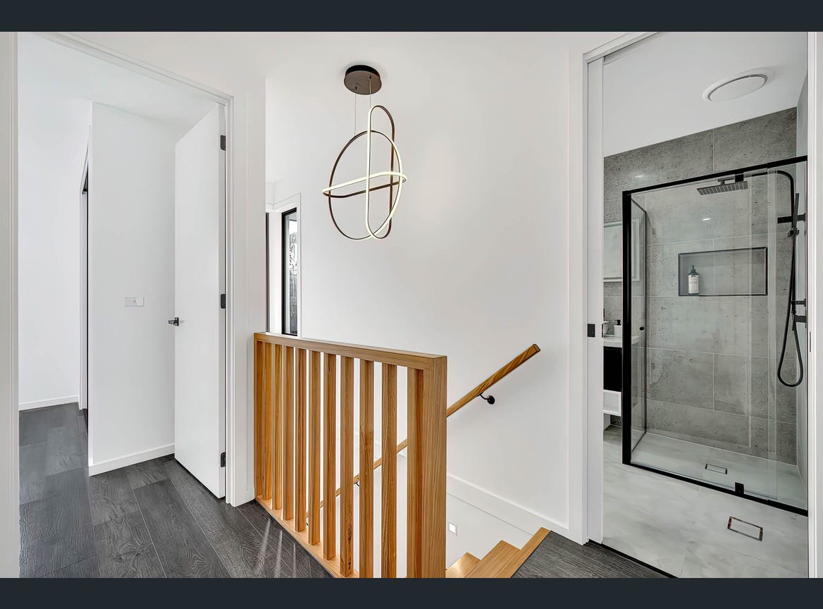 A bright hallway showcases modern design elements with a wooden staircase leading to the upper level. A stylish light fixture hangs from the ceiling, and a glass-enclosed shower can be seen through a nearby doorway, enhancing the contemporary feel of the space.