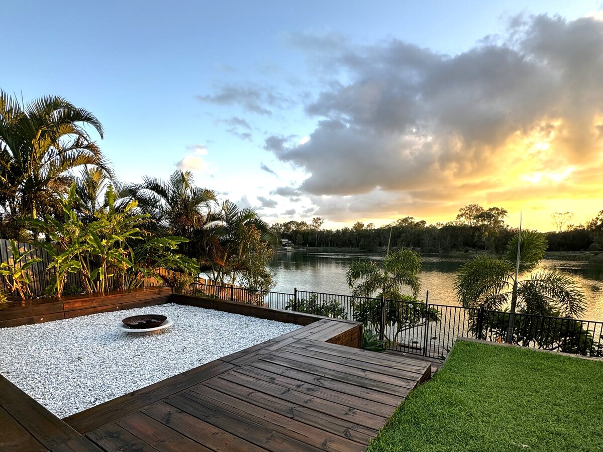 A wooden deck features a gravel area with a central firepit, surrounded by lush tropical plants. The scene is bathed in warm sunset light, with the serene waters of Lake Kalvert visible in the background, framed by trees and distant horizon.