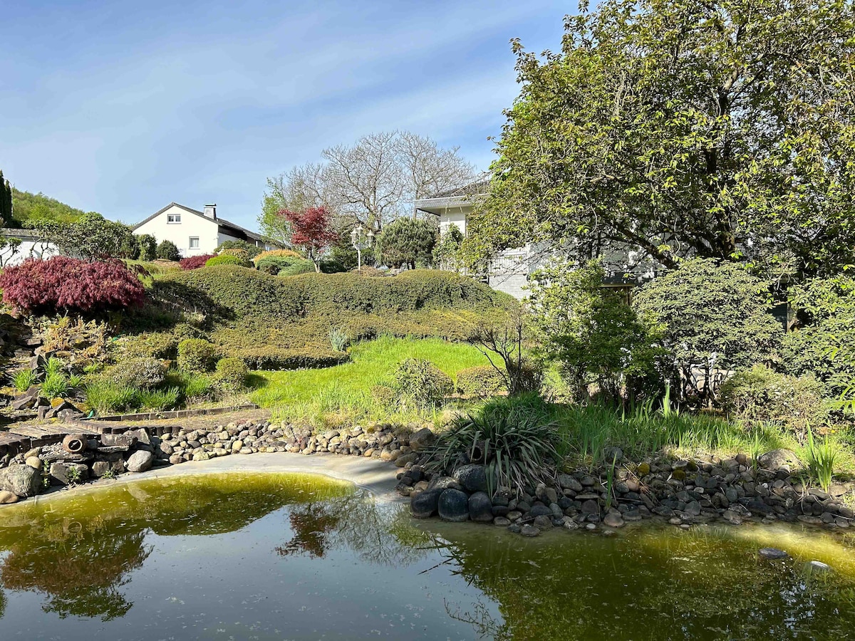 A peaceful garden is depicted, featuring a variety of plants and shrubs in shades of green and red. A small pond reflects the surrounding flora, while a gently sloping hillside is visible in the background. The area is marked by well-maintained paths and stone elements.