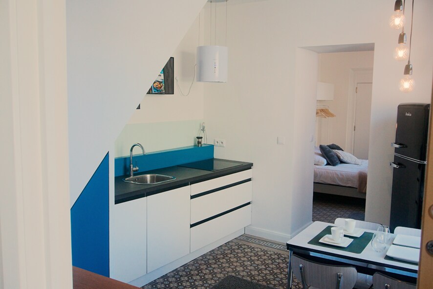 A contemporary kitchen area features a sleek countertop and a sink beneath a minimalist light fixture. The open layout connects to a cozy sleeping area visible in the background, showcasing a spacious bed and a light color palette.