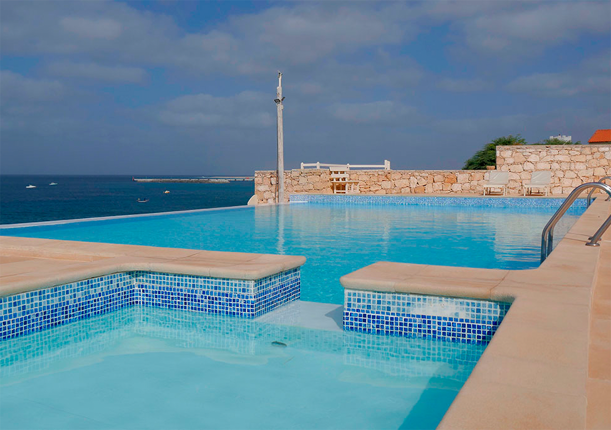 An infinity pool is visible, featuring crystal-clear water blending seamlessly with the ocean horizon. The pool is surrounded by a stone wall, with sun loungers placed along the edge. Gentle waves can be seen in the distance against a backdrop of a blue sky.