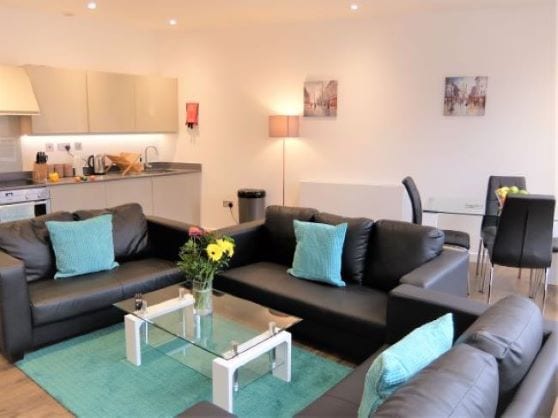 A modern living area features dark leather sofas arranged around a glass coffee table, which holds a vase of fresh flowers. A dining table with four chairs is positioned in the background, and a well-equipped kitchen is visible to the left.