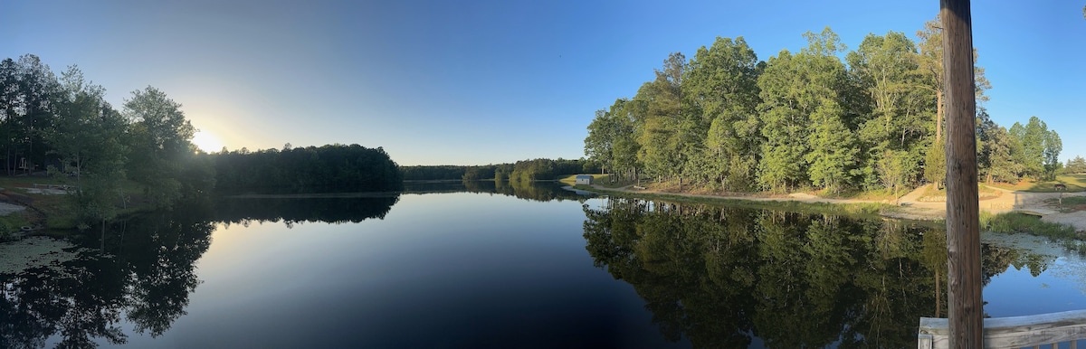 A panoramic view showcases a calm lake nestled among lush greenery. The still water reflects the vibrant blue sky and surrounding trees, creating a serene environment for relaxation and outdoor activities. A gentle slope leads to a sandy area on the right bank.