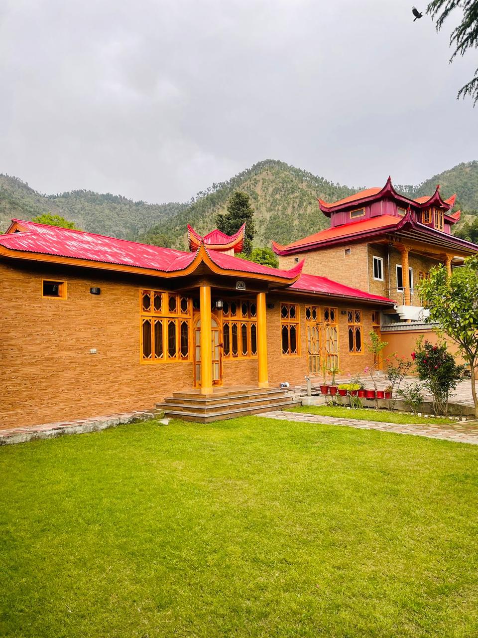 A charming wooden cottage featuring traditional architectural elements, set against a backdrop of rolling hills. The structure is characterized by vibrant red roofs and intricate window designs, surrounded by a well-maintained lawn that invites relaxation and outdoor enjoyment.
