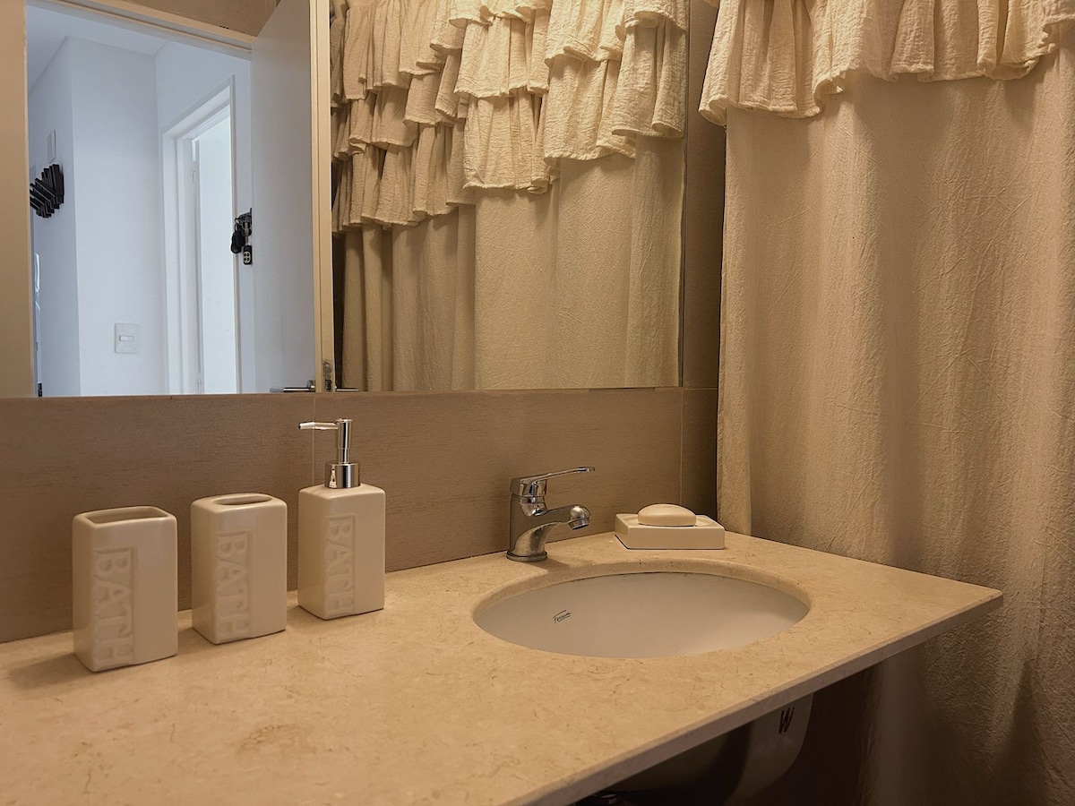 A clean and well-appointed bathroom features a light-colored stone countertop with matching dispensers and containers. The spacious mirror reflects soft lighting from above. A neutral-colored shower curtain adds warmth, while the overall design emphasizes simplicity and functionality.