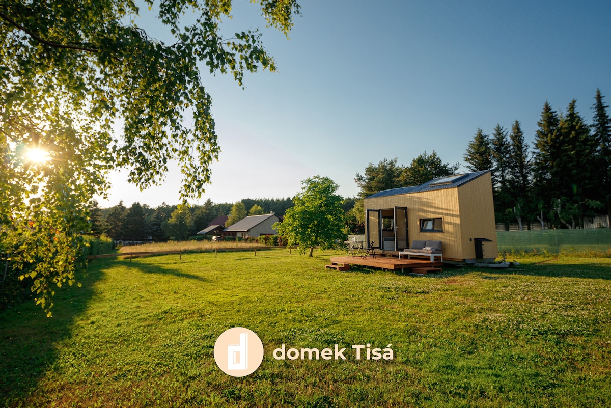 A cozy holiday accommodation is set in a green landscape, highlighted by a front deck and large windows. Sunlight filters through the trees, creating a warm atmosphere. The surrounding lawn is open and spacious, offering a serene environment.