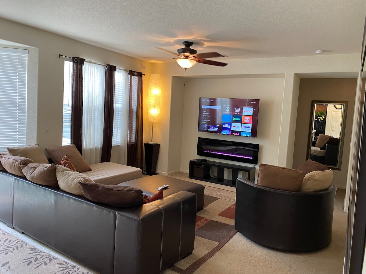 A spacious living area features a central sectional sofa with complementing chairs positioned around a coffee table. A large smart TV is mounted on the wall, with a soft glow from a floor lamp illuminating the room. Natural light filters in through the windows, adorned with light curtains.