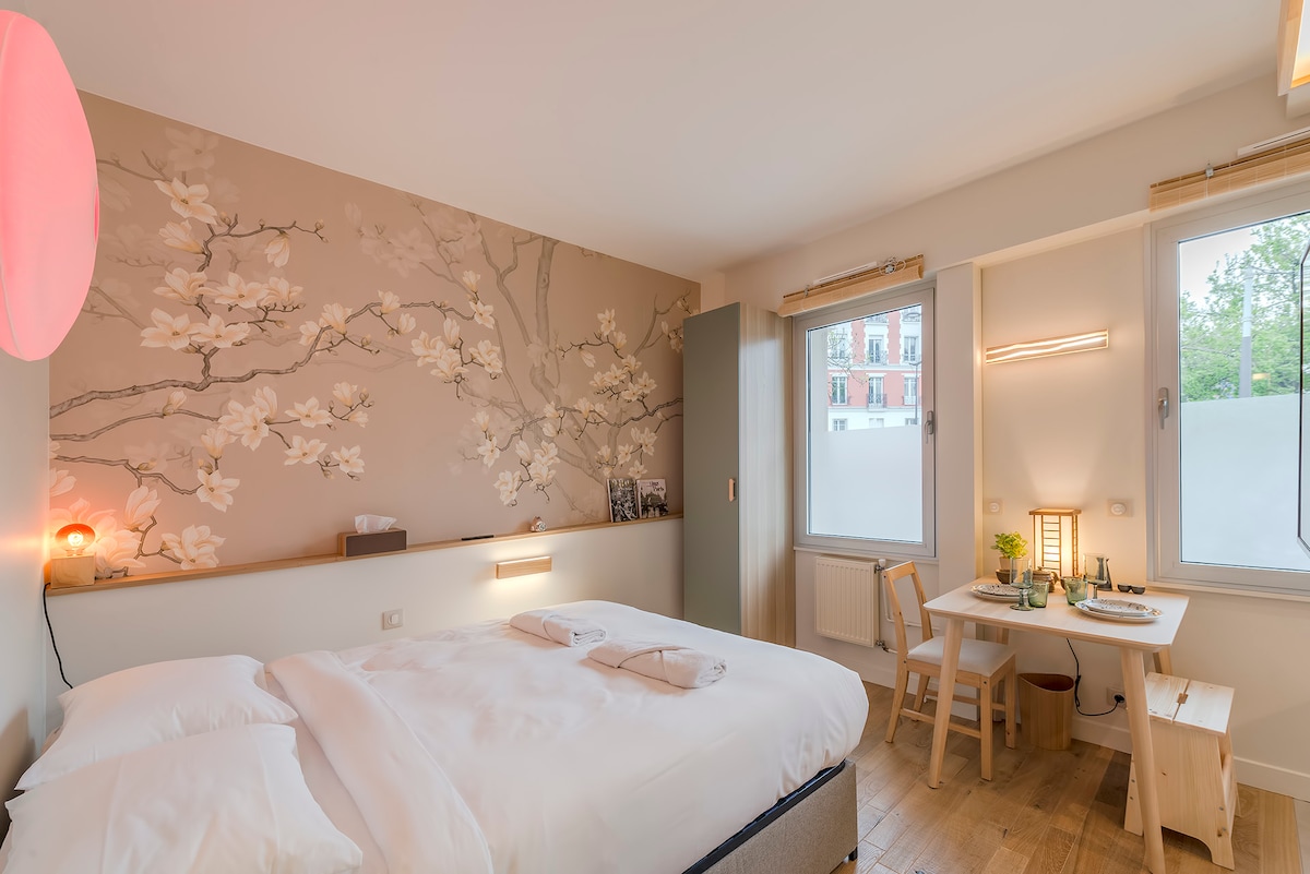 A spacious bedroom features a floral accent wall adorned with delicate white blossoms. A cozy queen-size bed with crisp linens is positioned opposite a small dining area with two chairs. Natural light streams through large windows, enhancing the calming atmosphere.