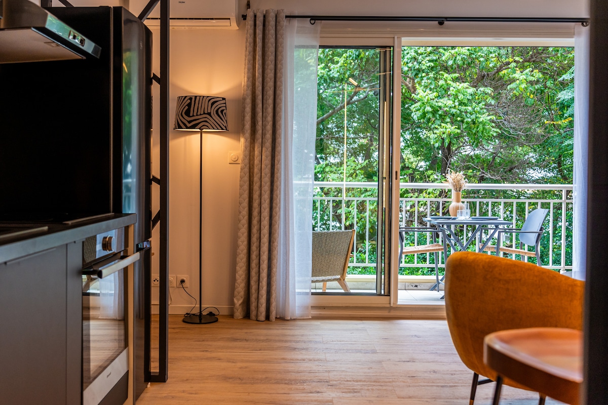 A living area features a modern kitchen setup with a sleek black cabinet. Natural light floods through sliding glass doors that open to a private balcony, where outdoor seating is arranged. Lush greenery is visible beyond the balcony, adding to the serene ambiance.