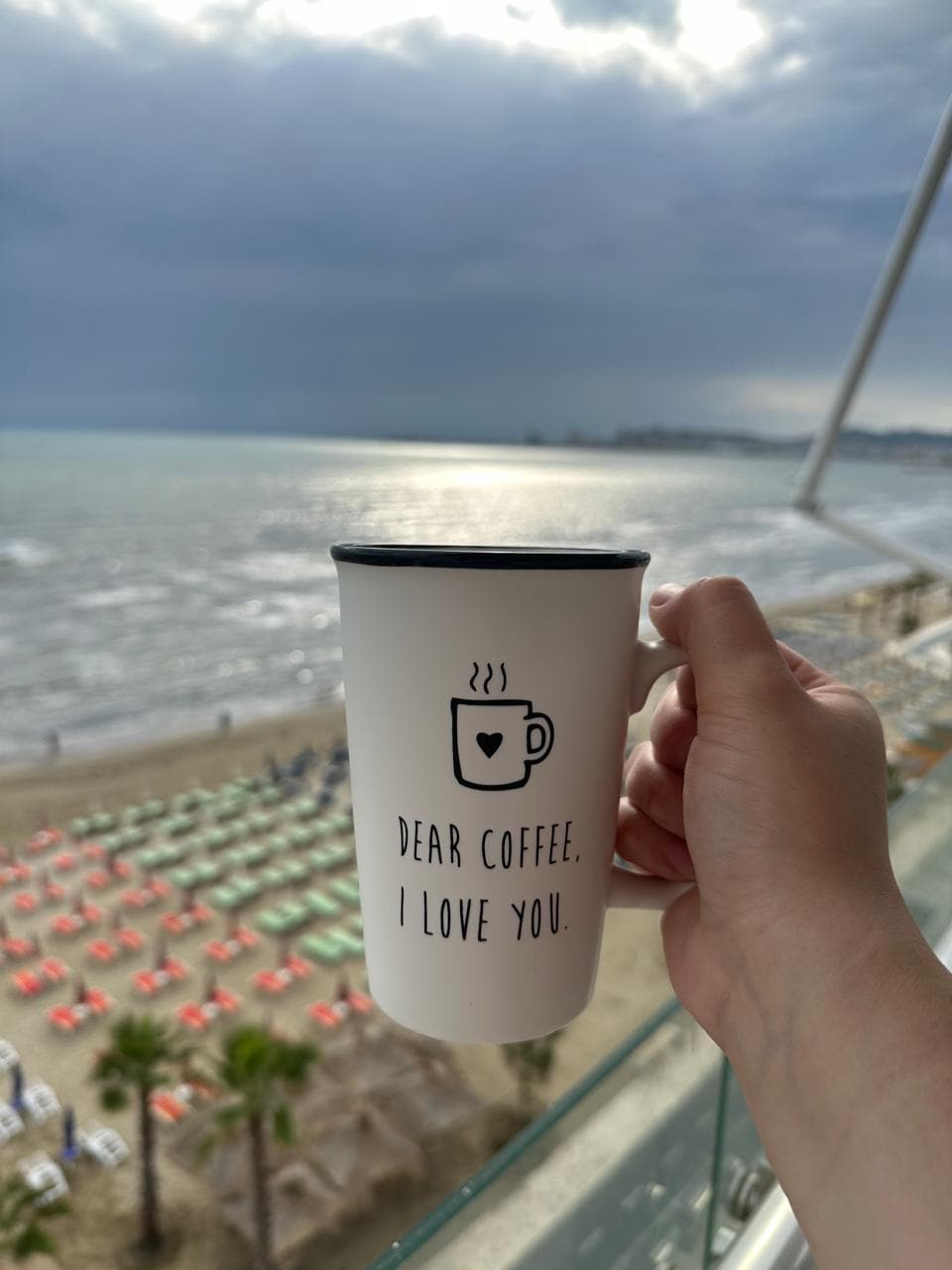 A hand holds a white coffee mug featuring the words 'DEAR COFFEE. I LOVE YOU.' against a backdrop of the sea. The view includes beach umbrellas and loungers in the sand, along with a cloudy sky reflecting over the water.