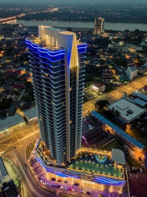An aerial view of a tall modern building showcases its sleek architecture and illuminated rooftop. Surrounding streets highlight the vibrant urban landscape, with a tranquil river visible in the background, offering a glimpse of the central location.