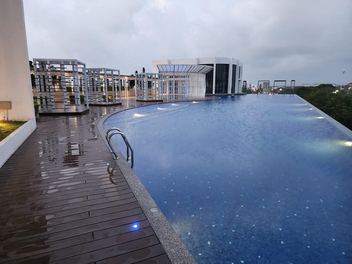 An open-air infinity pool extends toward the horizon, surrounded by sleek wooden decking. Soft lighting illuminates the space, enhancing the pool's tranquil blue water. Lush greenery is visible in the background, while modern seating areas provide spots for relaxation.