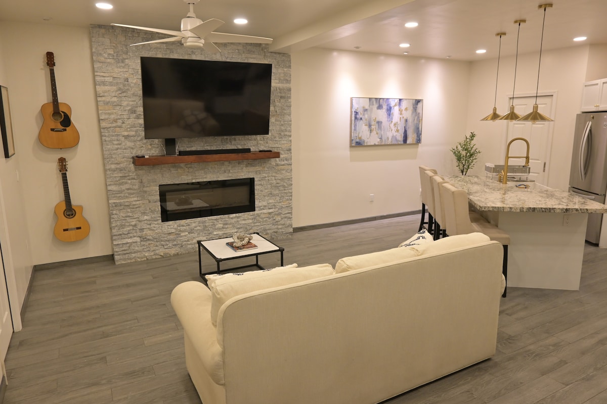 A light and airy living space features a comfortable cream-colored sofa facing a modern fireplace. Two acoustic guitars are displayed on the wall, and a mounted television provides entertainment. A marble kitchen island with four bar stools offers a gathering space, illuminated by stylish pendant lights.