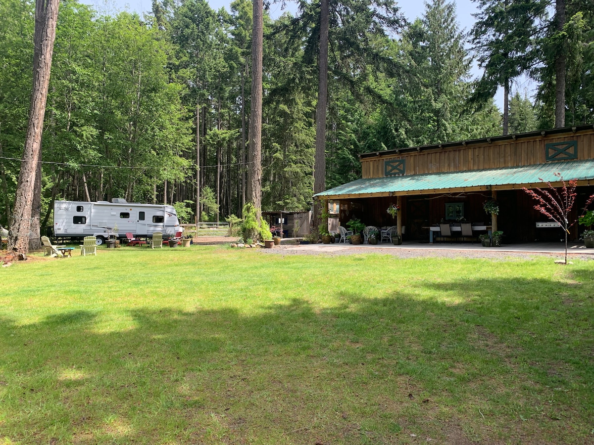 A spacious grassy area is framed by tall trees, leading to a large barn with a covered seating area. A silver trailer is parked nearby, providing additional accommodations. The natural surroundings offer a serene atmosphere, perfect for outdoor relaxation or gatherings.