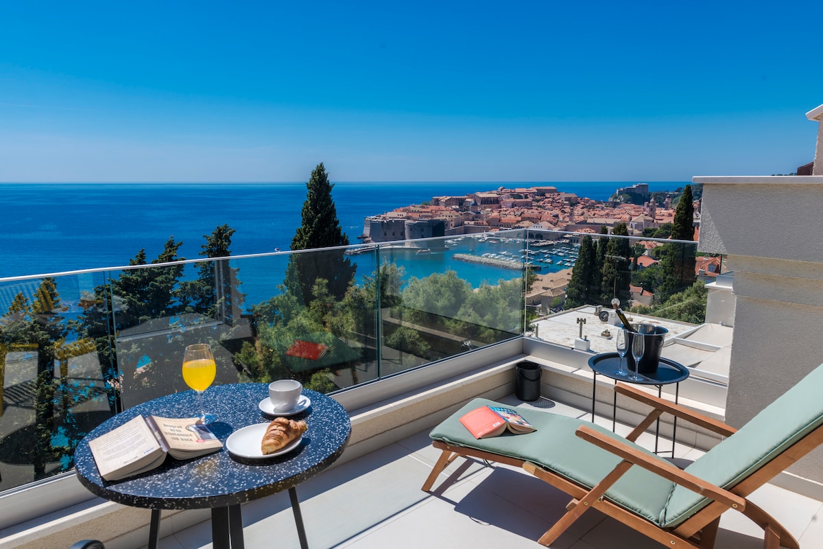 A sunlit balcony features a comfortable lounge chair and a small table set with a book, a croissant, and a glass of orange juice. Views of the sea and Old Town are visible in the background, separated by a glass railing that enhances the open atmosphere.