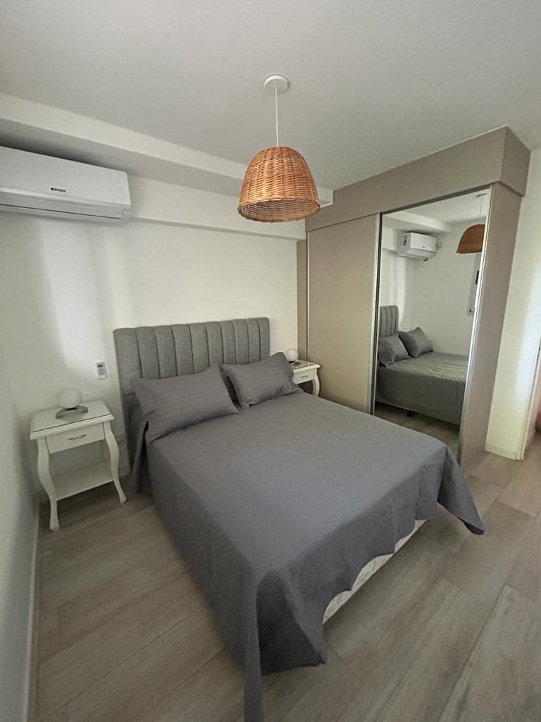 A well-appointed bedroom features a spacious bed with grey linens and matching pillows. A stylish rattan pendant light hangs above. Mirrored doors add depth and reflect natural light, while a bedside table with a lamp is positioned on one side of the bed.