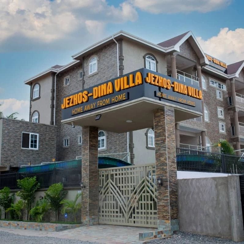 The entrance of Jezhos-Dina Villa is showcased, featuring a decorative stone gate with the name prominently displayed. Surrounding greenery adds a touch of nature, while the multi-story building stands in the background under a blue sky with soft clouds.