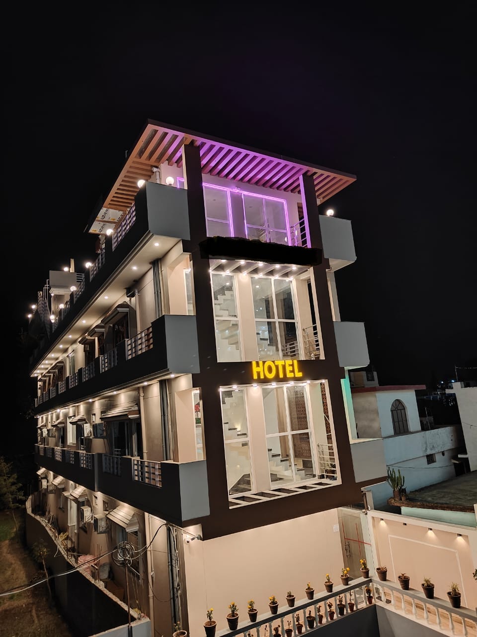 The hotel building is prominently displayed with modern architectural features and multiple floors. Illuminated signage reads 'HOTEL' on the exterior. Warm lighting accents the structure, with large windows providing an open and inviting look at night.