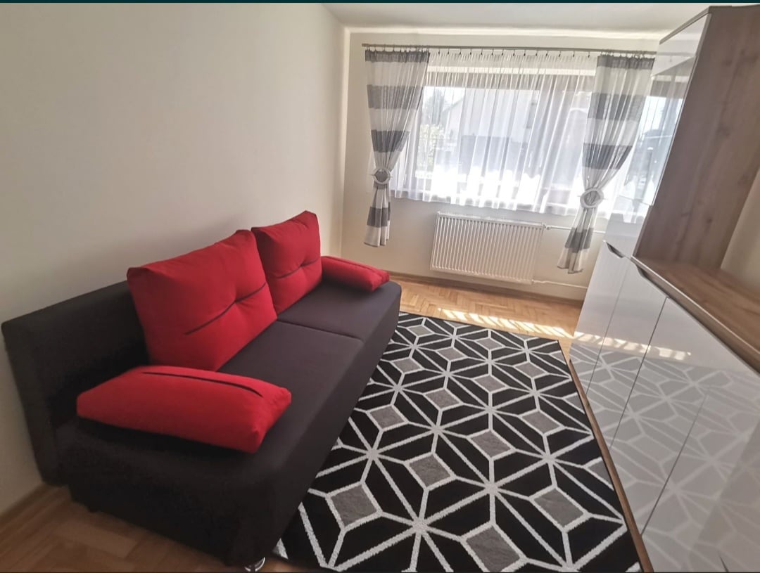 A modern living space is presented with a dark sofa accented by vibrant red pillows. A geometric patterned rug lies on the hardwood floor, complementing the room's furnishings. Light streams in through large windows adorned with sheer curtains, while a white storage unit is positioned against the wall.
