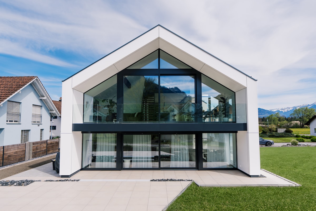 A modern luxury apartment is showcased with a striking triangular roof and large glass windows, allowing natural light to flood the interior. The clean white facade is complemented by a smooth stone pathway leading to a grassy area, creating a harmonious connection with the outdoors.