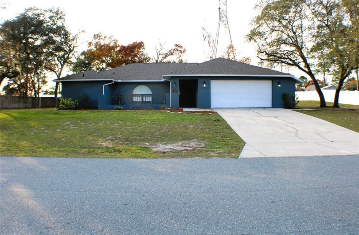The exterior of a single-story home is shown, featuring a blue façade and a front yard that is mostly grass. A long driveway leads to the attached garage, while trees provide shade along the property line.