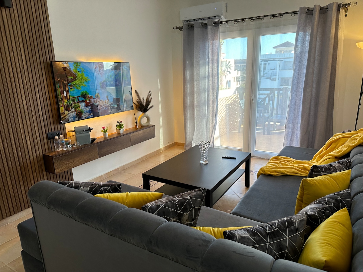 The living area features a comfortable gray sectional sofa adorned with yellow cushions, paired with a low coffee table. A wall-mounted television is visible above a sleek wooden shelf, while glass doors lead to a balcony, allowing natural light to fill the space.