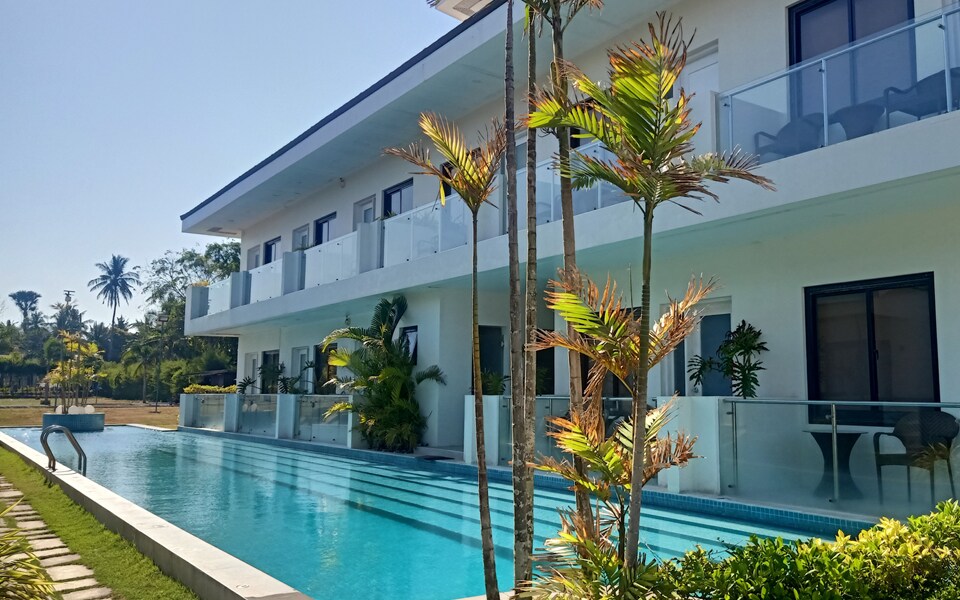 The guesthouse is depicted with a modern design, featuring a sleek swimming pool extending alongside its exterior. Lush greenery and palm plants surround the property, while spacious balconies are visible on the second floor, providing an inviting atmosphere for relaxation.