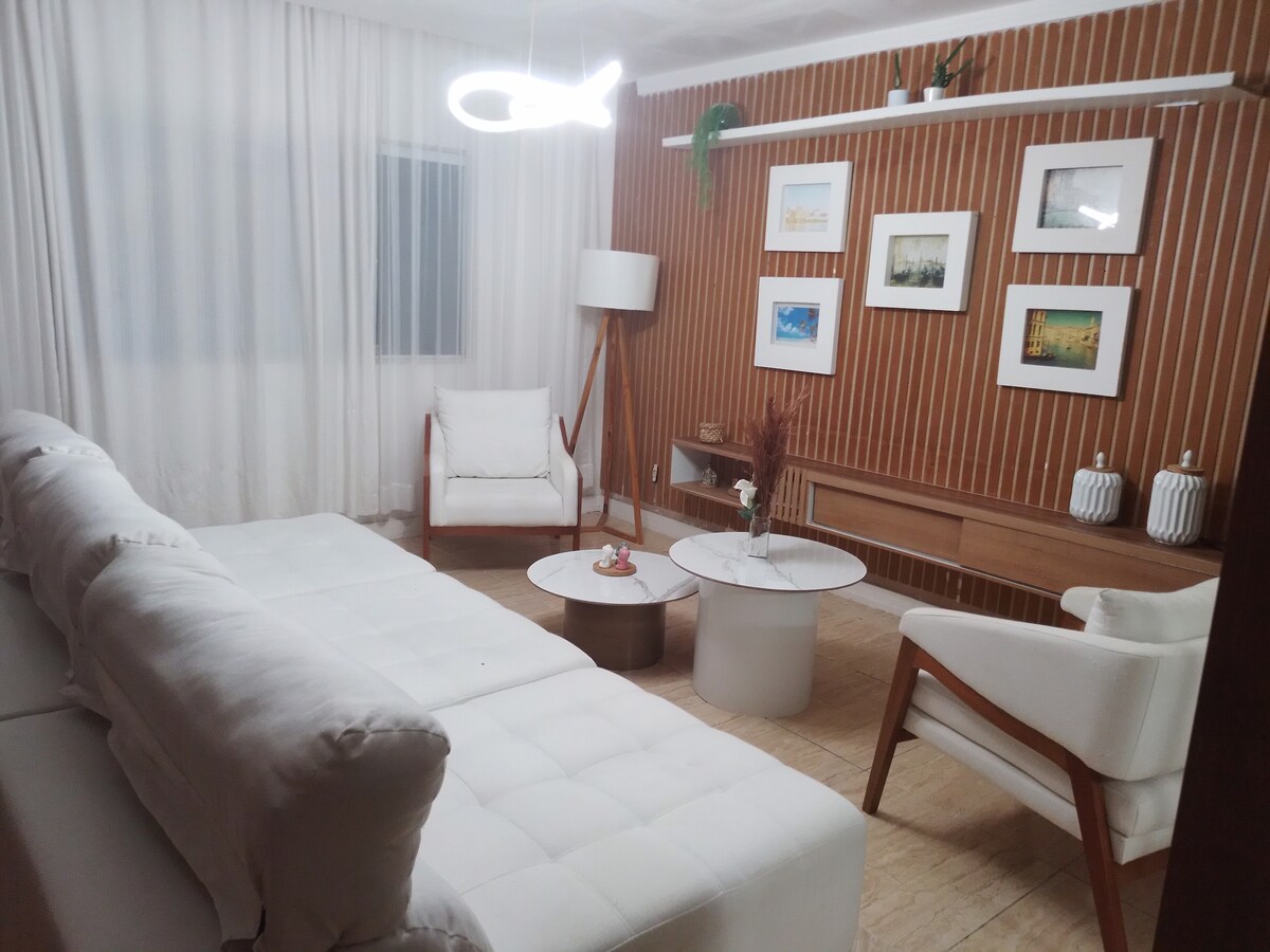 The living area features a modern white sectional sofa positioned alongside a minimalist round coffee table. A pair of white chairs complements the space, with soft curtains draping over a window. Framed artworks and decorative vases adorn the wood-paneled wall, enhancing the room's inviting atmosphere.