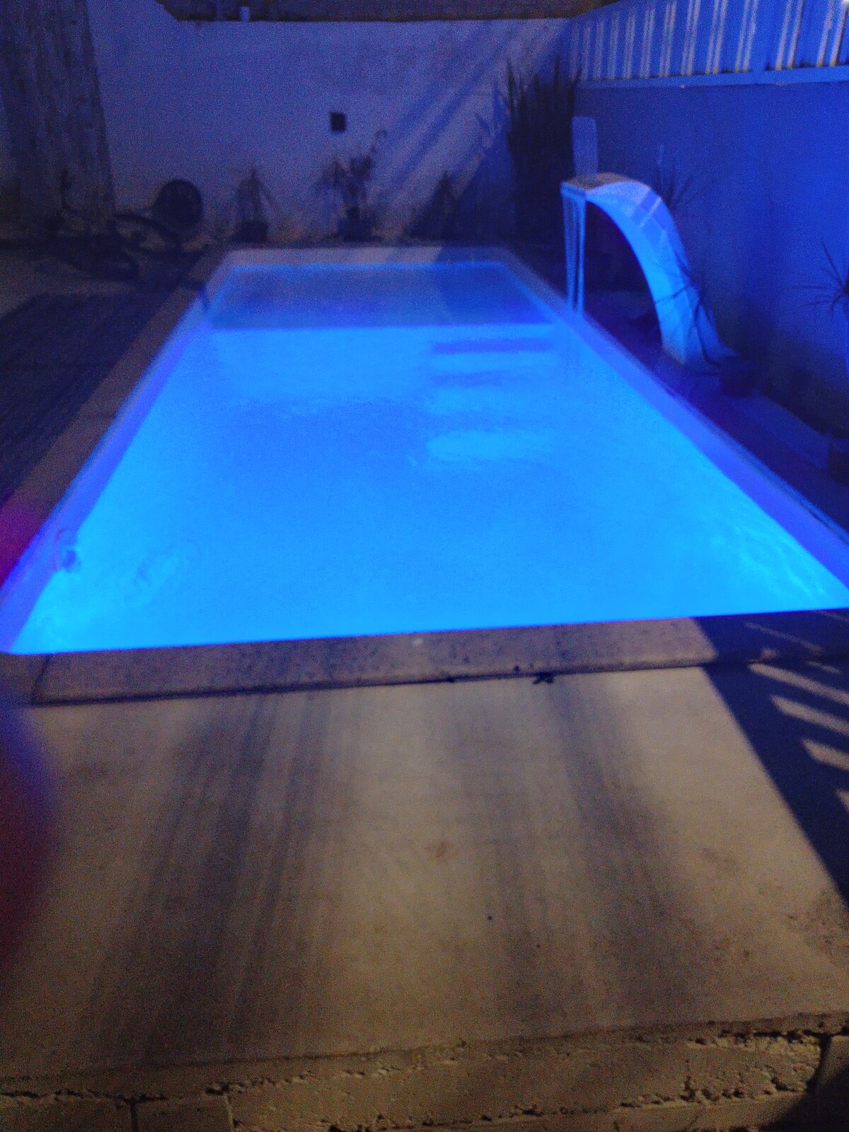 A tranquil pool is illuminated with soft blue lighting, creating a calming ambiance. The pool features a smooth surface and a gentle waterfall, surrounded by minimalistic decor and potted plants, offering a serene space for relaxation.