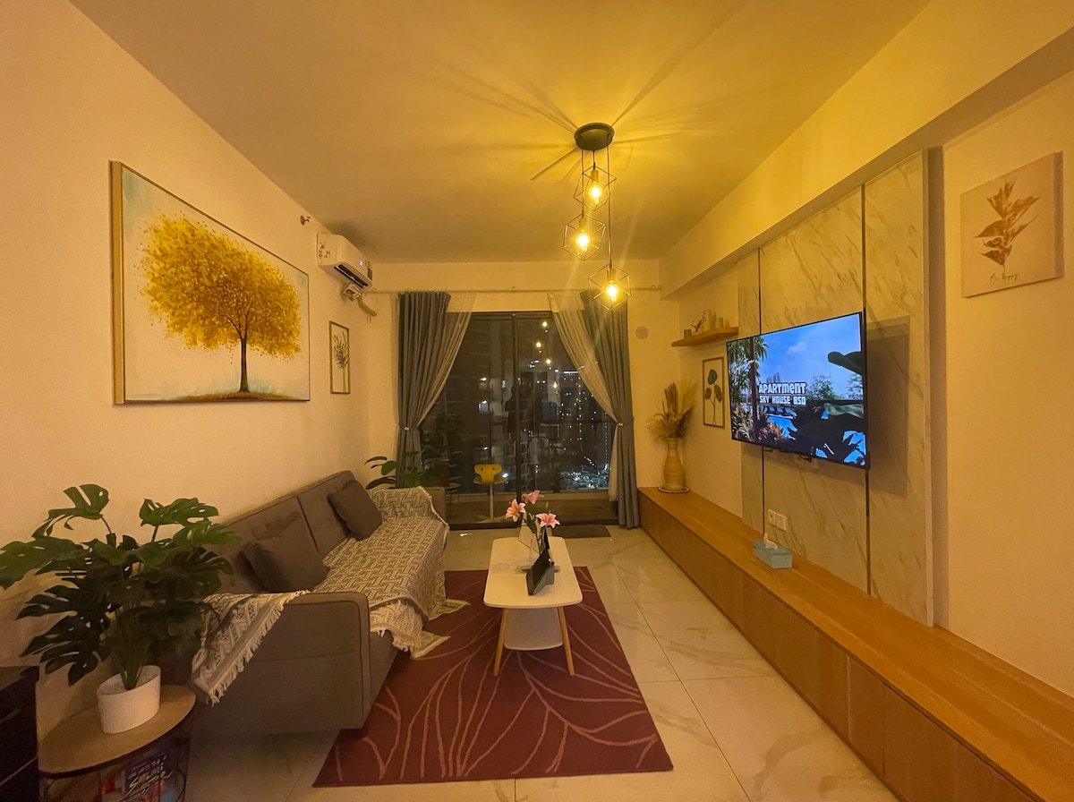 A spacious living room is highlighted by a modern design. A comfortable sofa is positioned beside a coffee table on a patterned rug. A large television is mounted against a light-toned feature wall, with soft lighting creating a relaxed atmosphere. A window offers a view of the city.