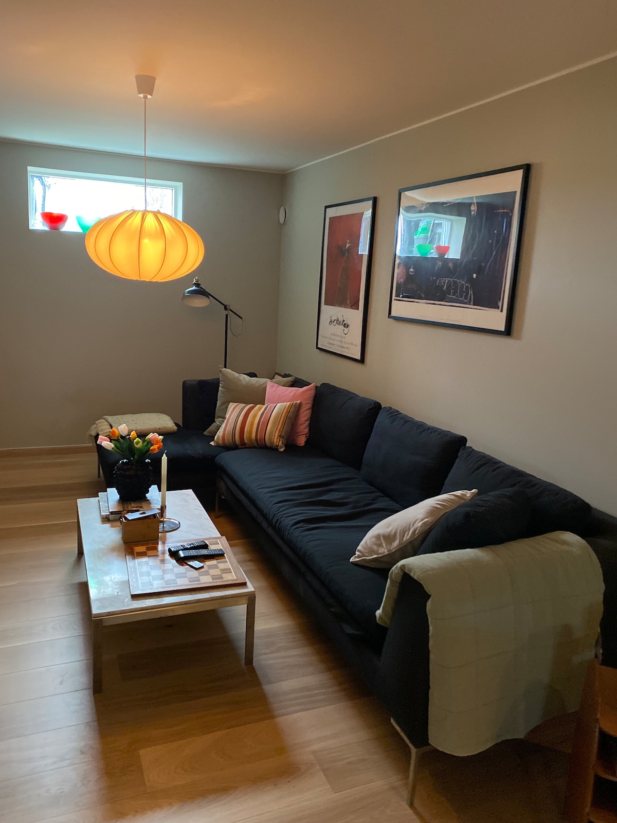 A comfortable corner features a dark sofa adorned with cushions, facing a light wooden coffee table. Two art pieces are displayed on the wall, and a pendant light hangs above the seating area. A small window allows natural light to brighten the space.