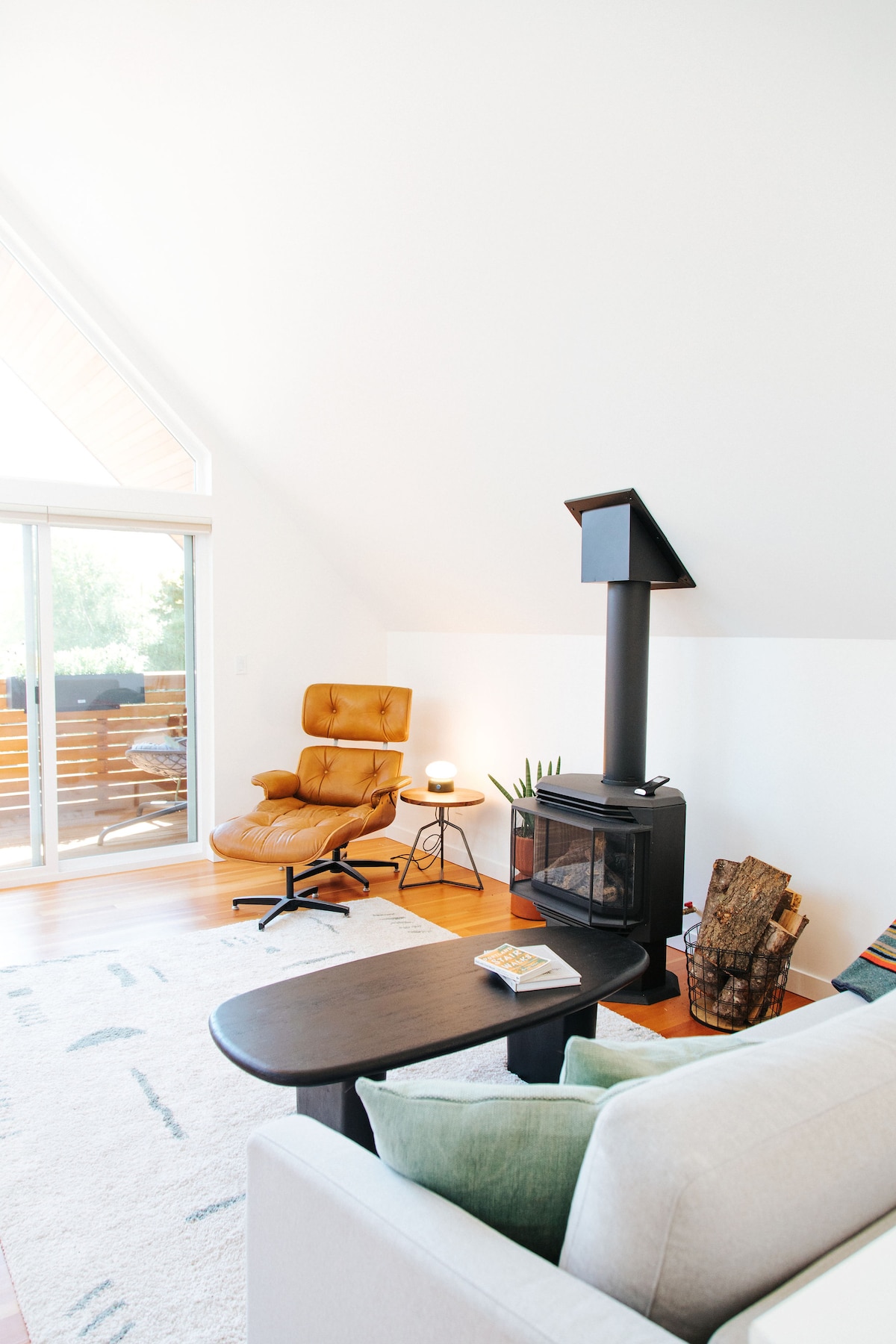 A modern living space is highlighted by a leather armchair beside a small side table. A wood-burning stove and a neutral-toned sofa add to the coziness. Large glass doors allow natural light and provide access to a private deck.