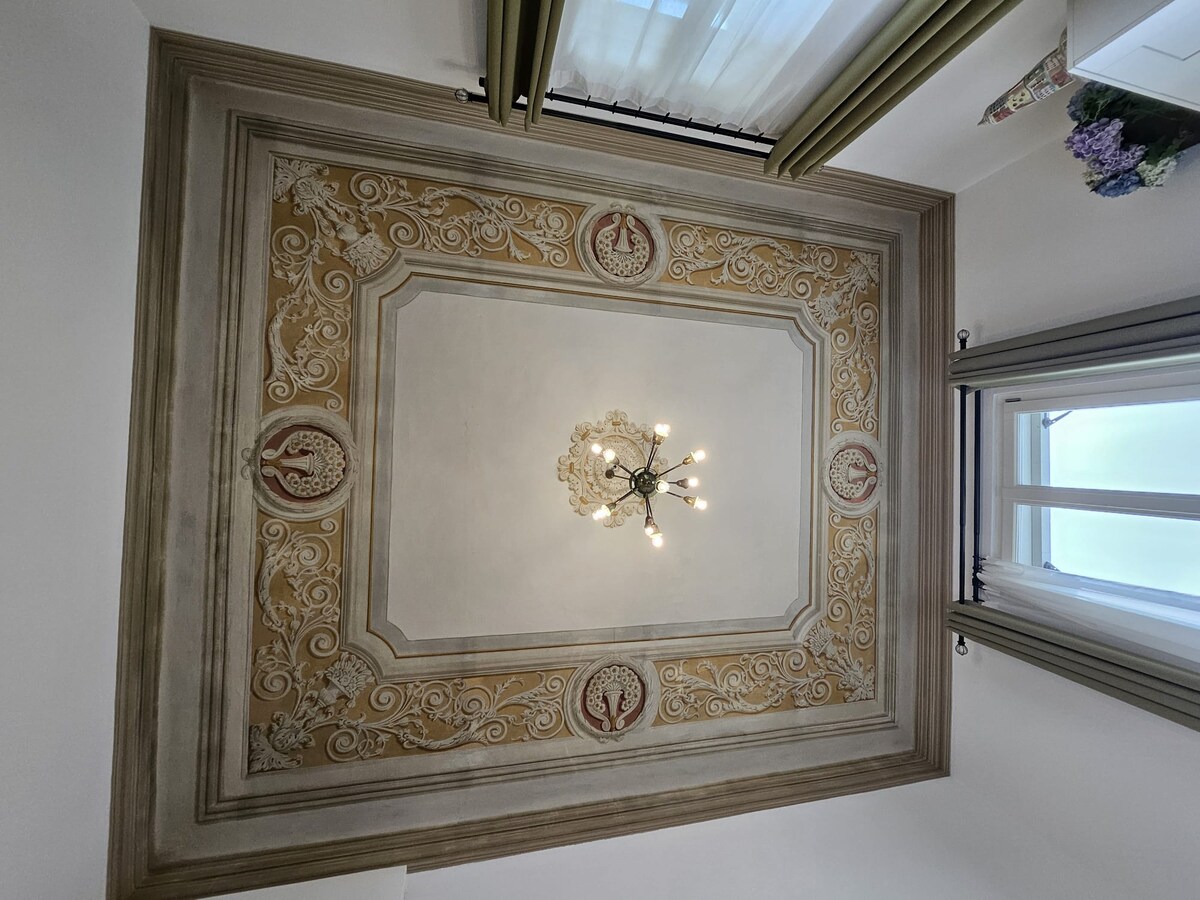 A detailed ceiling showcases intricate frescoes framed by a decorative border. A stylish chandelier hangs at the center, providing soft illumination. Natural light filters in through the adjacent window, enhancing the elegance of the space.
