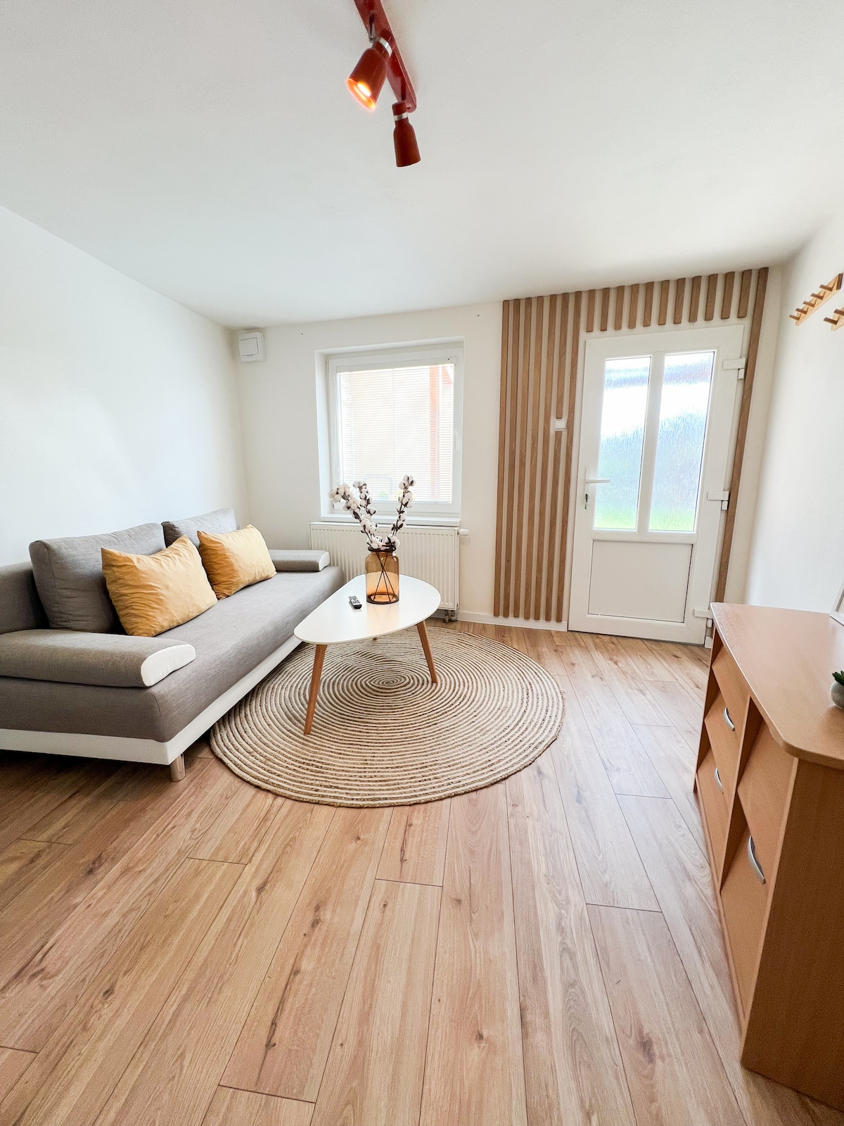 A light-filled living area includes a gray sofa with yellow throw pillows positioned beside a round coffee table. A decorative vase with neutral flowers complements the space. A wooden cabinet and a door leading outside are visible, enhancing the room's cozy atmosphere.