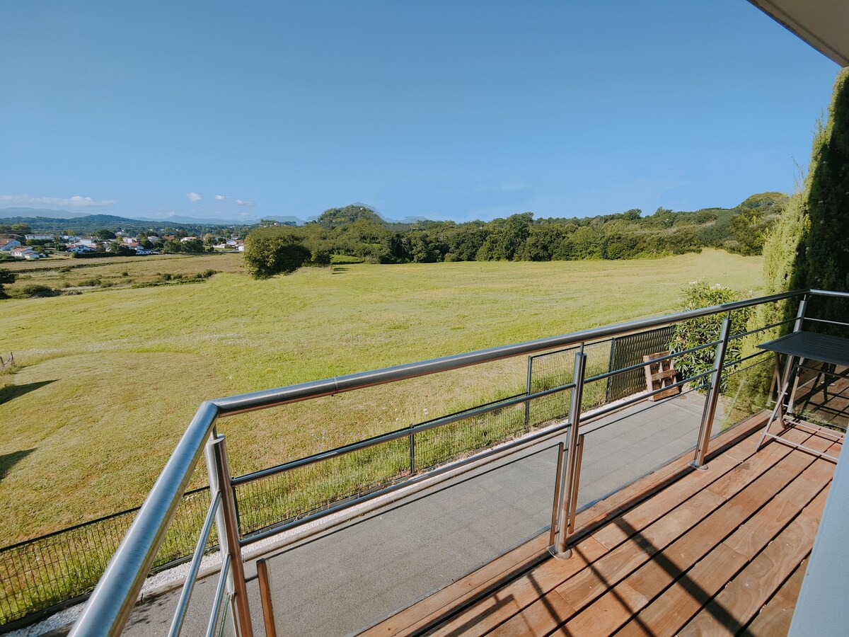 A spacious balcony is shown, featuring a railing that overlooks a vast expanse of greenery. The landscape extends towards distant hills under a clear blue sky, providing an open and serene atmosphere.