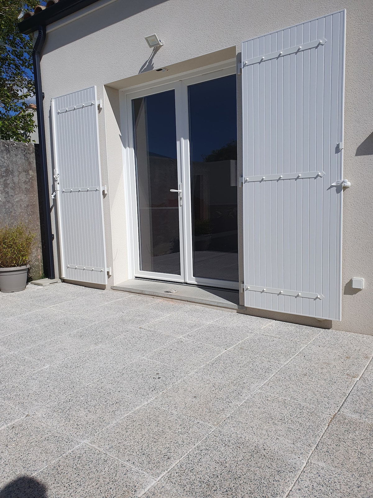 A set of double glass doors stands framed by white shutters, leading to a private outdoor area. The grey stone patio extends from the doors, offering a clean and modern appearance. Soft daylight highlights the space, while greenery is visible in the background.