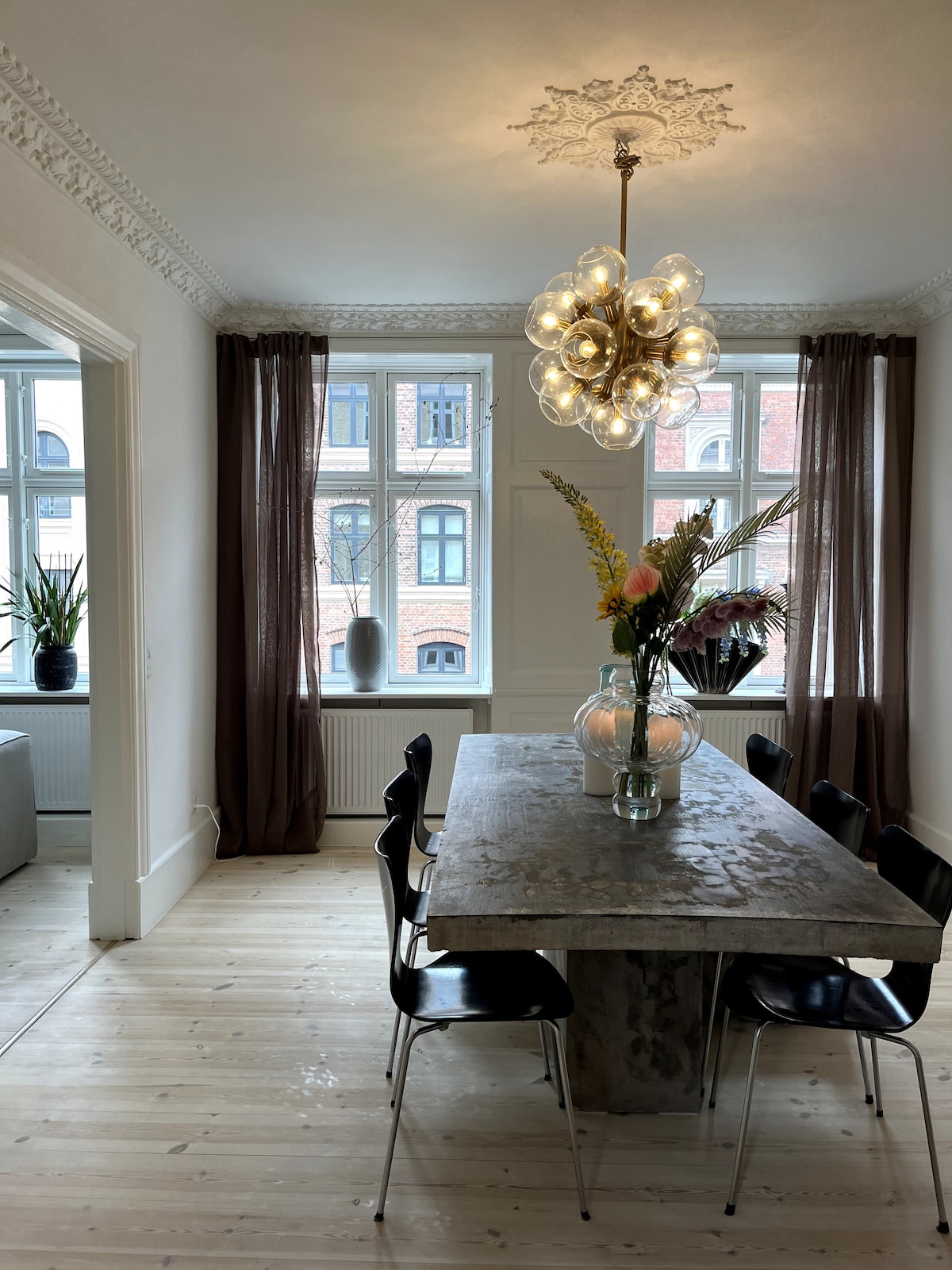A dining area features a large stone table surrounded by black chairs. Large windows allow natural light to fill the room, while light curtains adorn the sides. A decorative light fixture hangs from the ceiling, and a flower arrangement adds color to the table.