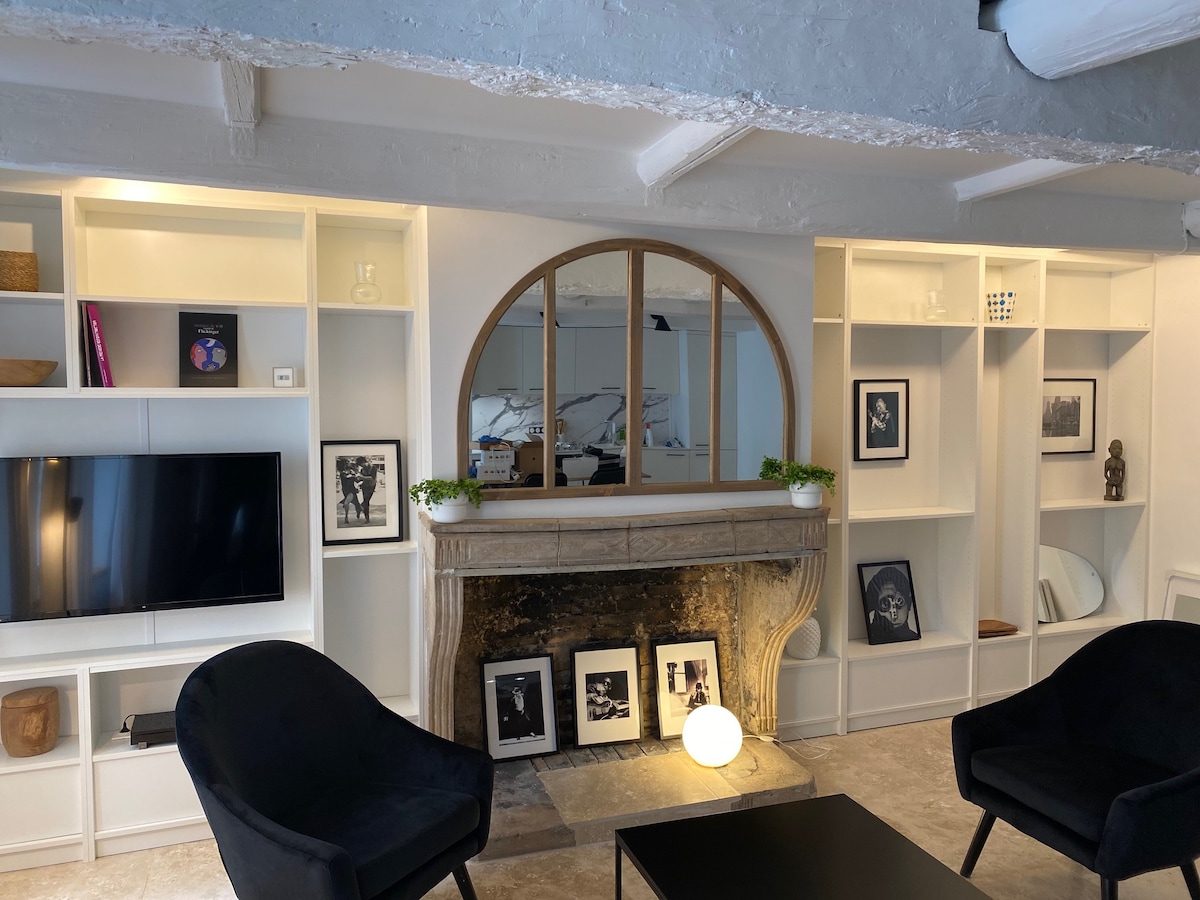 A bright living area is presented with a modern fireplace framed by a curved mirror. Black chairs are arranged around a low coffee table, while a television is mounted on the wall across from the seating. Shelving displays various decorative items and framed photographs.