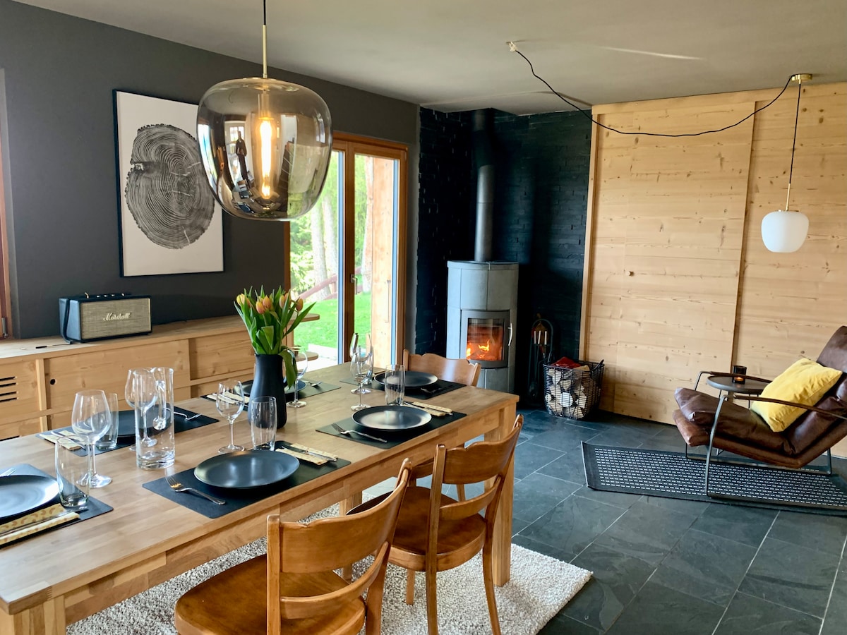 A dining area is showcased with a large wooden table set for six, accompanied by wooden chairs. A modern pendant light hangs overhead, and a cozy fireplace is visible in the background. Natural light enters through large windows, illuminating the space.