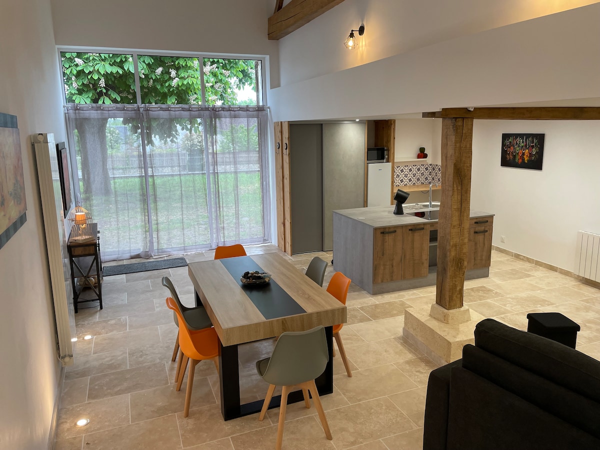 An open living area features a wooden dining table surrounded by colorful chairs. A modern kitchen is visible in the background, equipped with sleek appliances. Large windows allow natural light to fill the space, while a view of greenery is visible through sheer curtains.