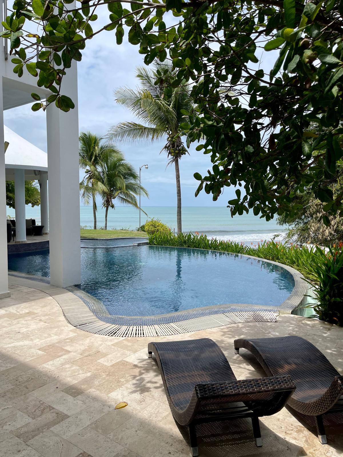 A serene outdoor area features a swimming pool with a smooth edge, surrounded by lush greenery. Two lounge chairs are positioned near the pool, offering views of the ocean and palm trees swaying gently in the breeze. Natural light brightens the tranquil setting.