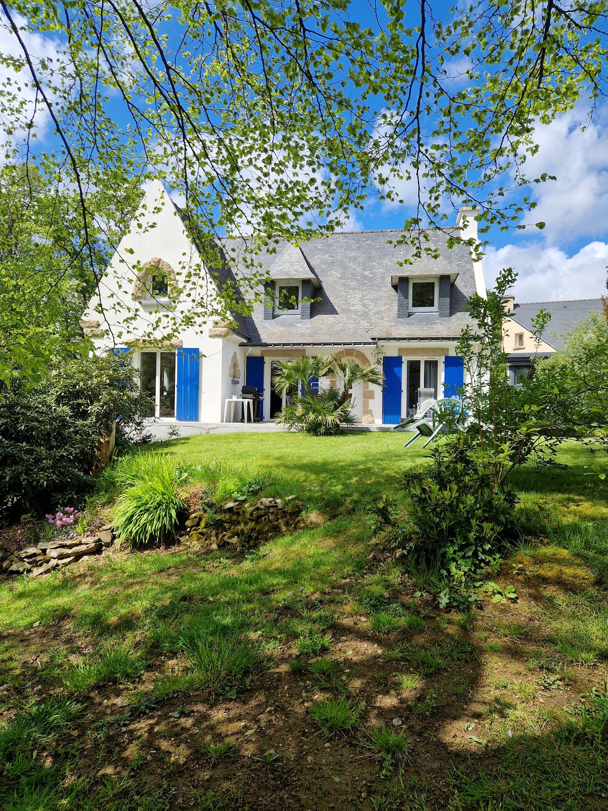 A charming house is nestled among greenery, featuring light-colored walls and blue shutters. Large windows allow natural light to illuminate the interior. A patio area with outdoor furniture is visible, surrounded by lush grass and a variety of plants, creating a peaceful setting.