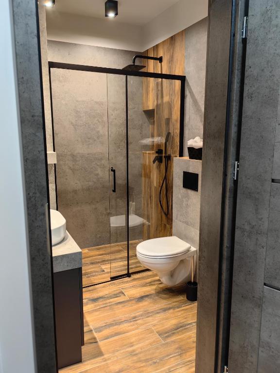 A modern bathroom features a spacious glass shower enclosure with a sleek black frame. Natural wood accents complement the concrete walls. A white sink is paired with a dark cabinet, while a toilet is positioned neatly beside it.