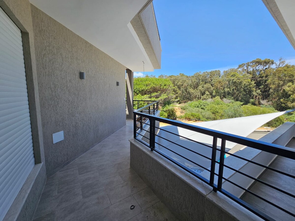A spacious balcony is shown, featuring a smooth tiled floor and a sleek railing. Lush greenery is visible beyond, providing a serene view. The walls are neutral-toned, and ample natural light brightens the area, highlighted by a clear blue sky.