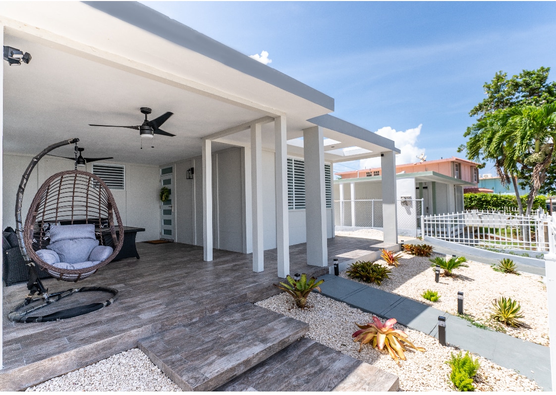A covered outdoor seating area features a hanging chair alongside a wooden deck. Tropical plants are arranged around the gravel landscape, and a ceiling fan provides ventilation. The bright exterior complements the clear sky and greenery in the background.