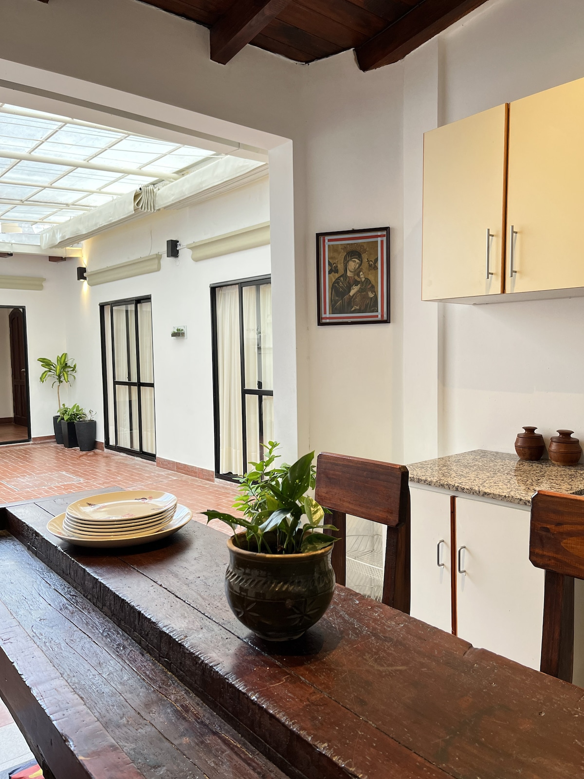 A modern kitchen space is highlighted by a polished wooden bar with plates, a potted plant, and granite countertops. Light floods the area through a skylight, illuminating the adjacent dining space and decorative artwork on the wall.