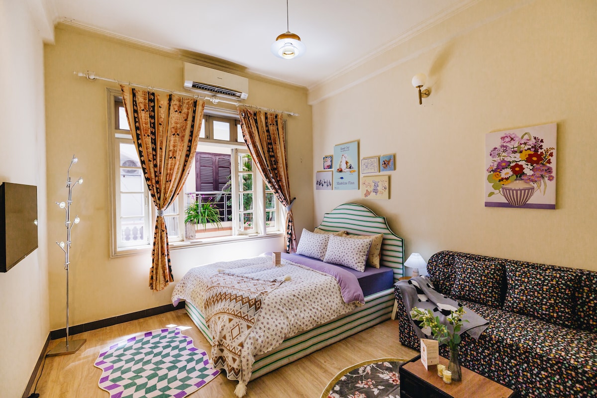 A calm bedroom setting includes a large bed adorned with patterned bedding and decorative cushions. A small sofa is positioned nearby, with vibrant floral decorations displayed on the wall. Natural light flows through the window, with soft curtains framing the view.