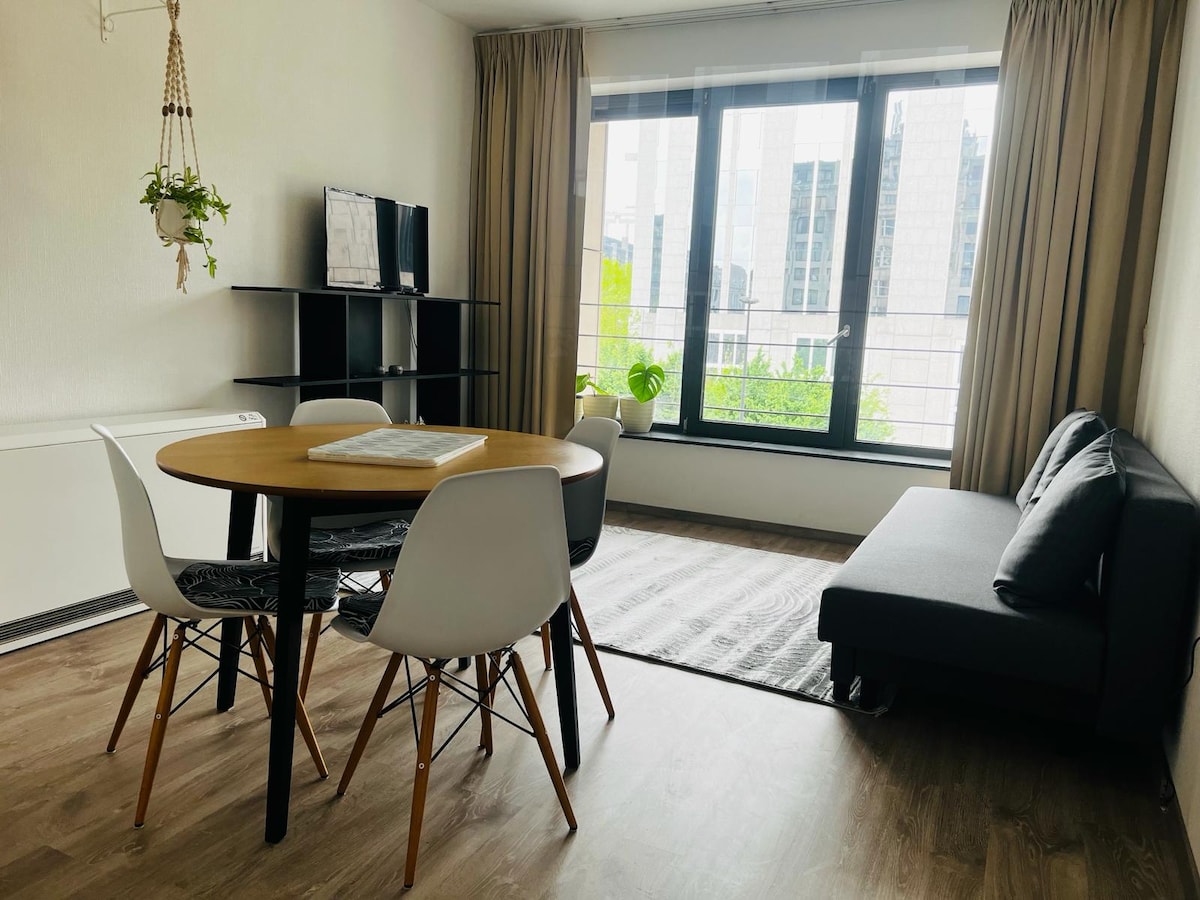 A bright living area features a round dining table surrounded by four chairs, complemented by a sleek black shelf and a flat-screen television. Large windows provide a view of the cityscape, with natural light illuminating the space and plants adding a touch of greenery.